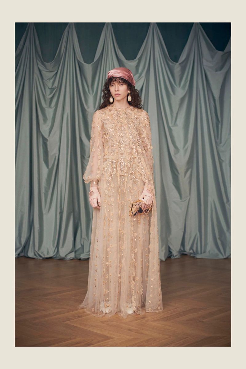 Woman in a light pink gown and head wrap poses against a draped backdrop. Long flowing dress, earrings, and neutral wood floor visible.