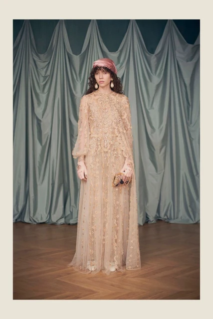 Woman in a light pink gown and head wrap poses against a draped backdrop. Long flowing dress, earrings, and neutral wood floor visible.