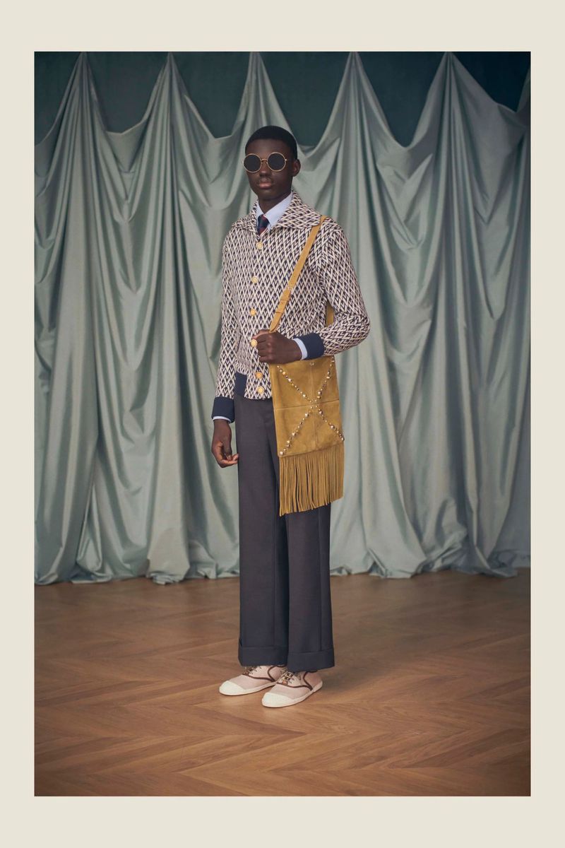 Fashion shoot features a slender Black male model wearing sunglasses and patterned jacket, standing indoors with a tan suede bag.