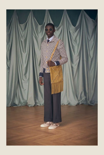 Fashion shoot features a slender Black male model wearing sunglasses and patterned jacket, standing indoors with a tan suede bag.