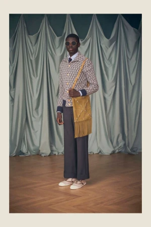 Fashion shoot features a slender Black male model wearing sunglasses and patterned jacket, standing indoors with a tan suede bag.