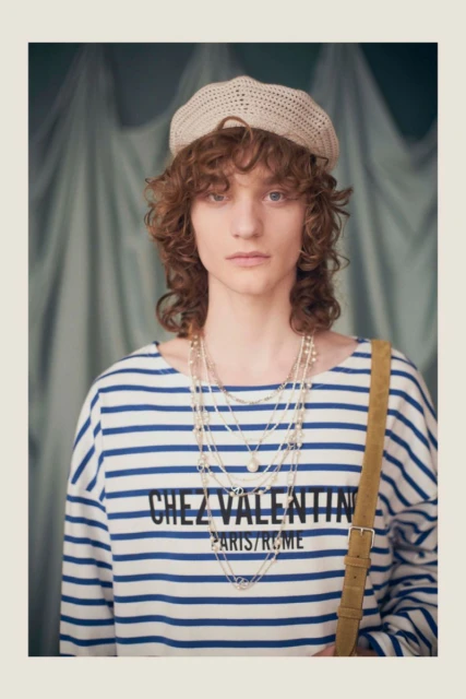 Adult fashion model wearing striped top, beret, and layered necklaces against a light blue backdrop in a medium portrait shot.