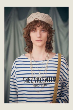 Adult fashion model wearing striped top, beret, and layered necklaces against a light blue backdrop in a medium portrait shot.