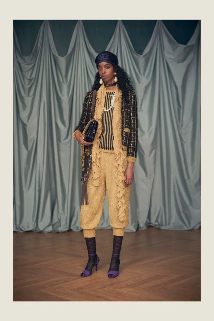 Fashionable Black woman in a black-and-gold jacket, matching pants, and turban poses in a studio with a dark teal curtain backdrop.