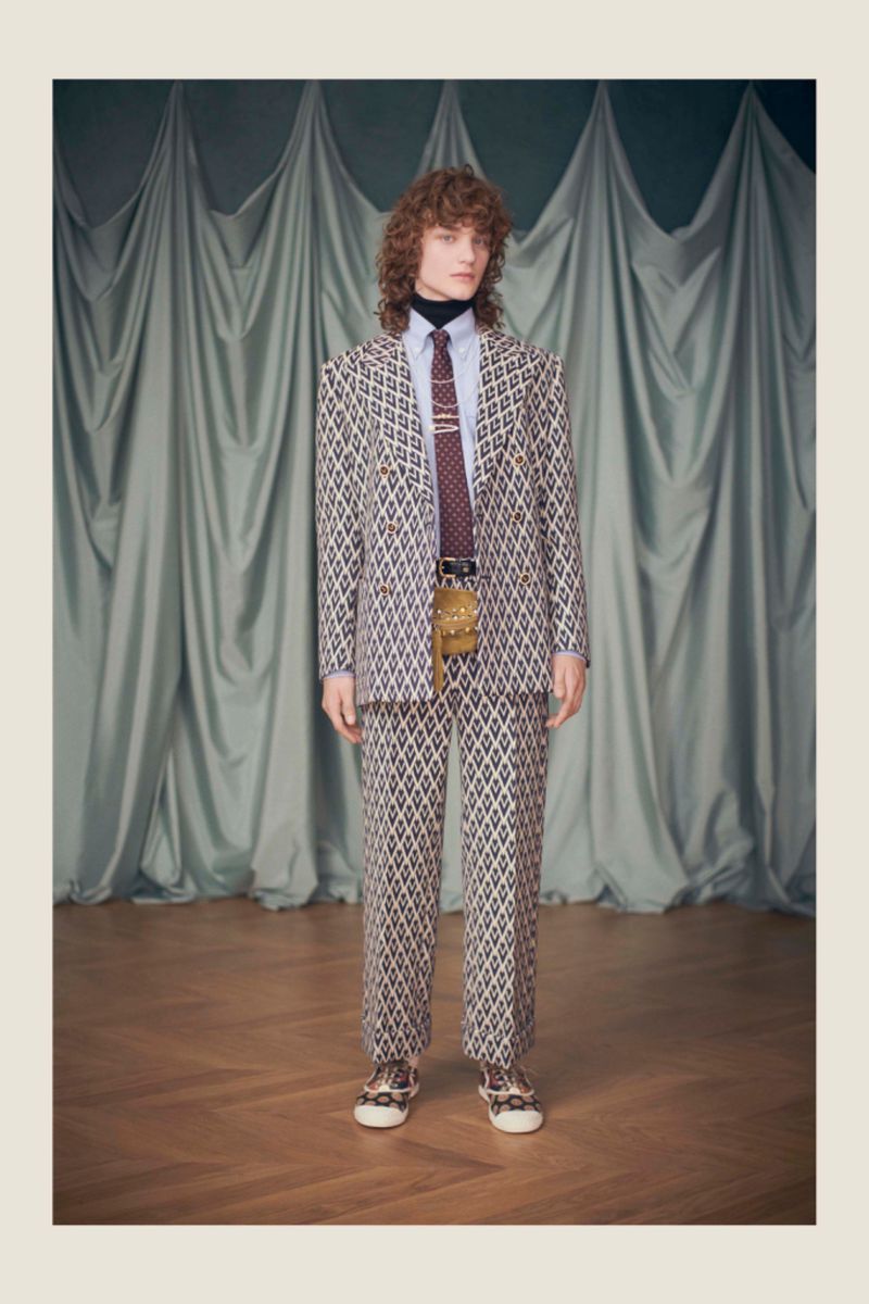 Full-length portrait of a male model wearing a diamond-patterned suit, turtleneck, and tie, posing indoors in front of a draped curtain.
