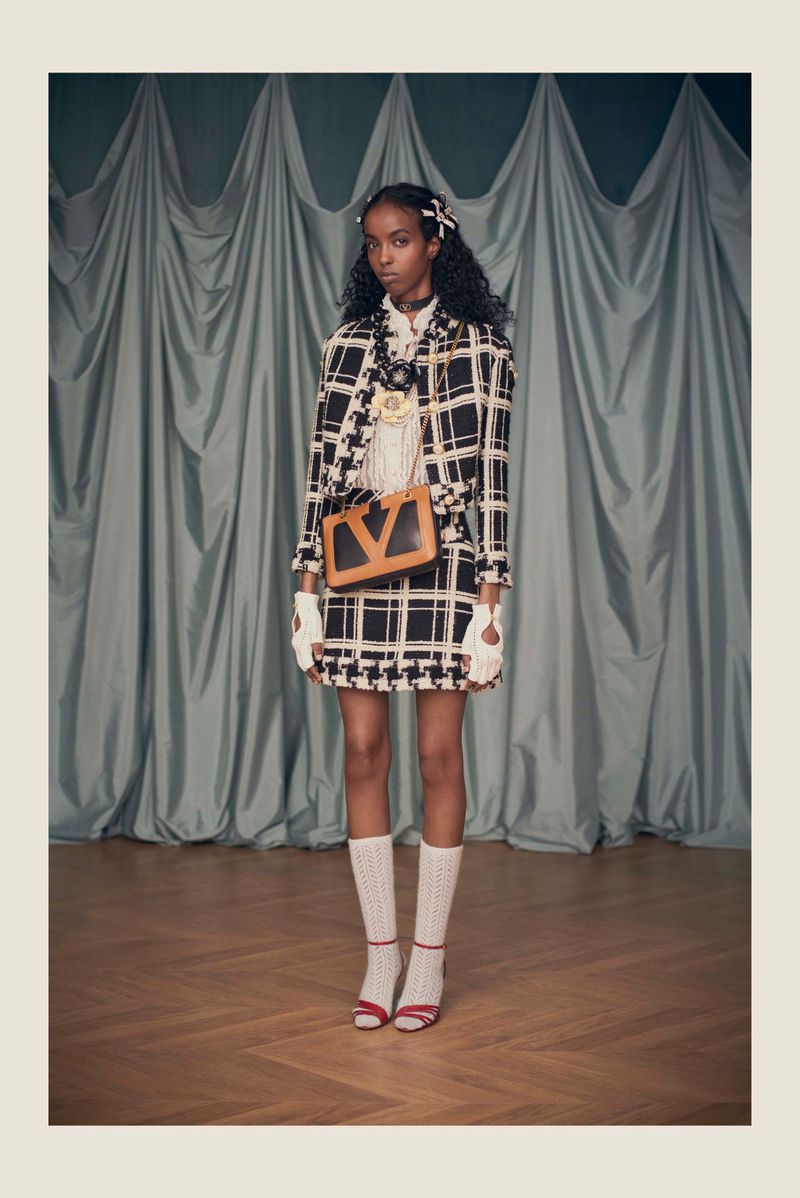 Fashion model in a black-and-white plaid skirt suit, with a light-brown handbag and white socks, poses against a green curtain backdrop.