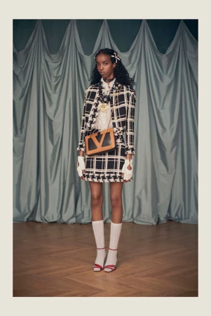 Fashion model in a black-and-white plaid skirt suit, with a light-brown handbag and white socks, poses against a green curtain backdrop.
