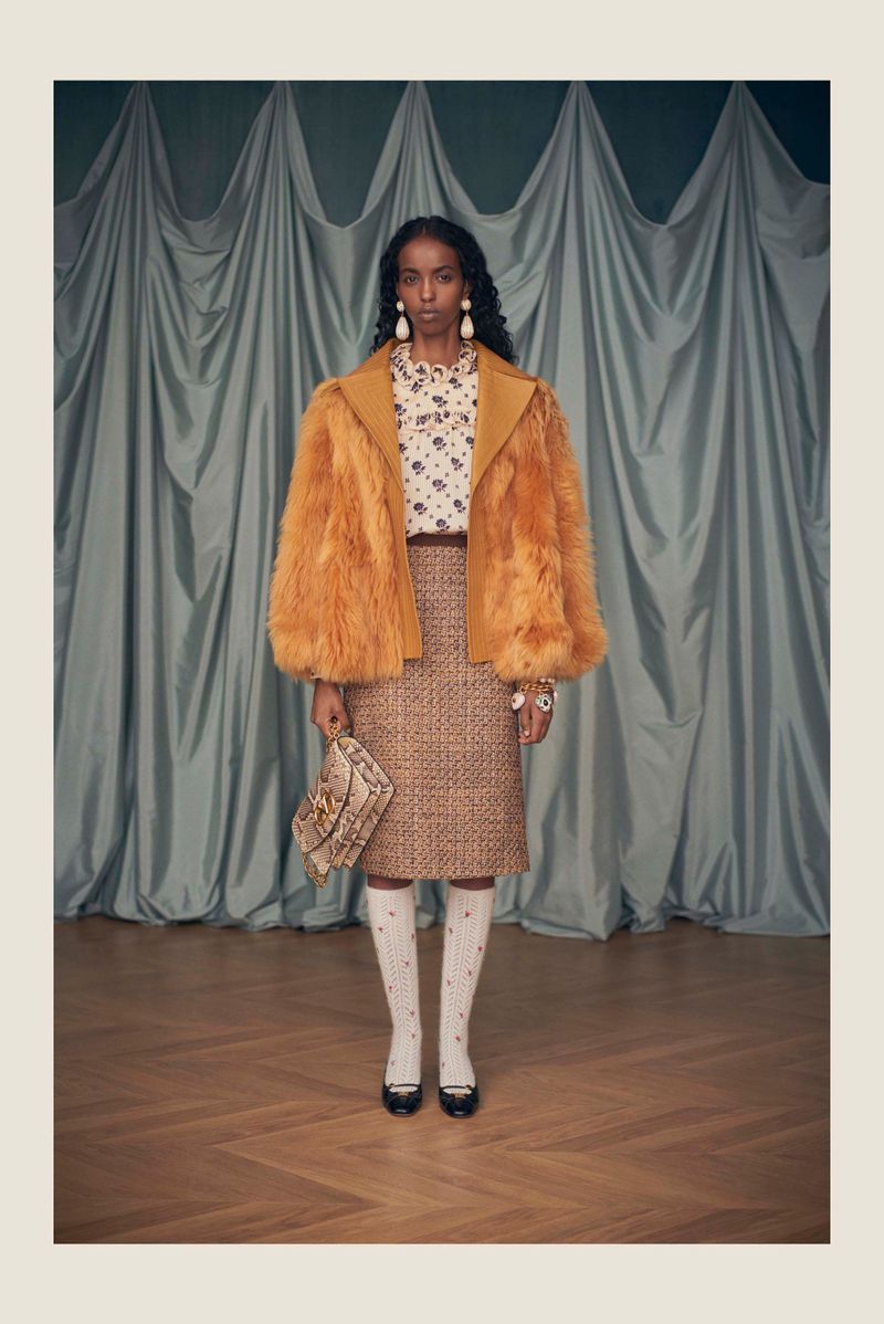 A dark-skinned woman poses in an orange faux-fur jacket with a patterned blouse and skirt, paired with knee socks and black shoes.