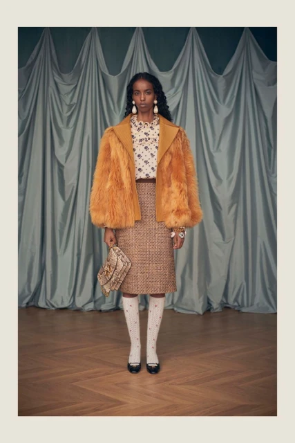 A dark-skinned woman poses in an orange faux-fur jacket with a patterned blouse and skirt, paired with knee socks and black shoes.