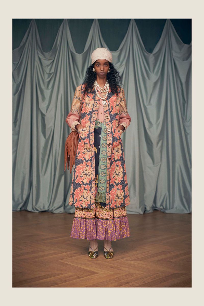 A woman in a floral coat stands in front of a curtain. She wears a hat, jewelry, and a patterned skirt, with a fringe purse over her shoulder.