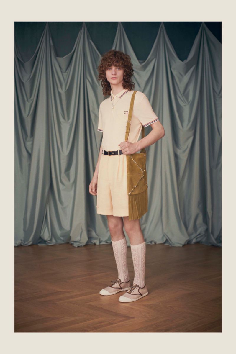 A young adult male poses in a studio wearing a polo shirt, shorts, long socks, and shoes, carrying a studded shoulder bag in a fashion shot.