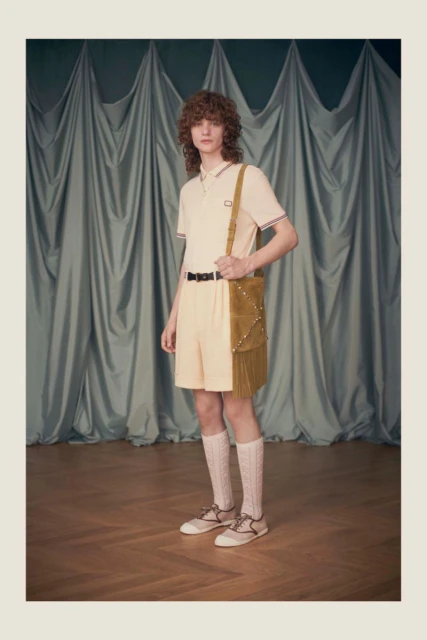 A young adult male poses in a studio wearing a polo shirt, shorts, long socks, and shoes, carrying a studded shoulder bag in a fashion shot.