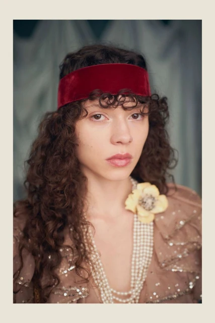 Portrait of a woman with curly hair and a red velvet headband, wearing a beige sequined dress and layered pearl necklaces indoors.