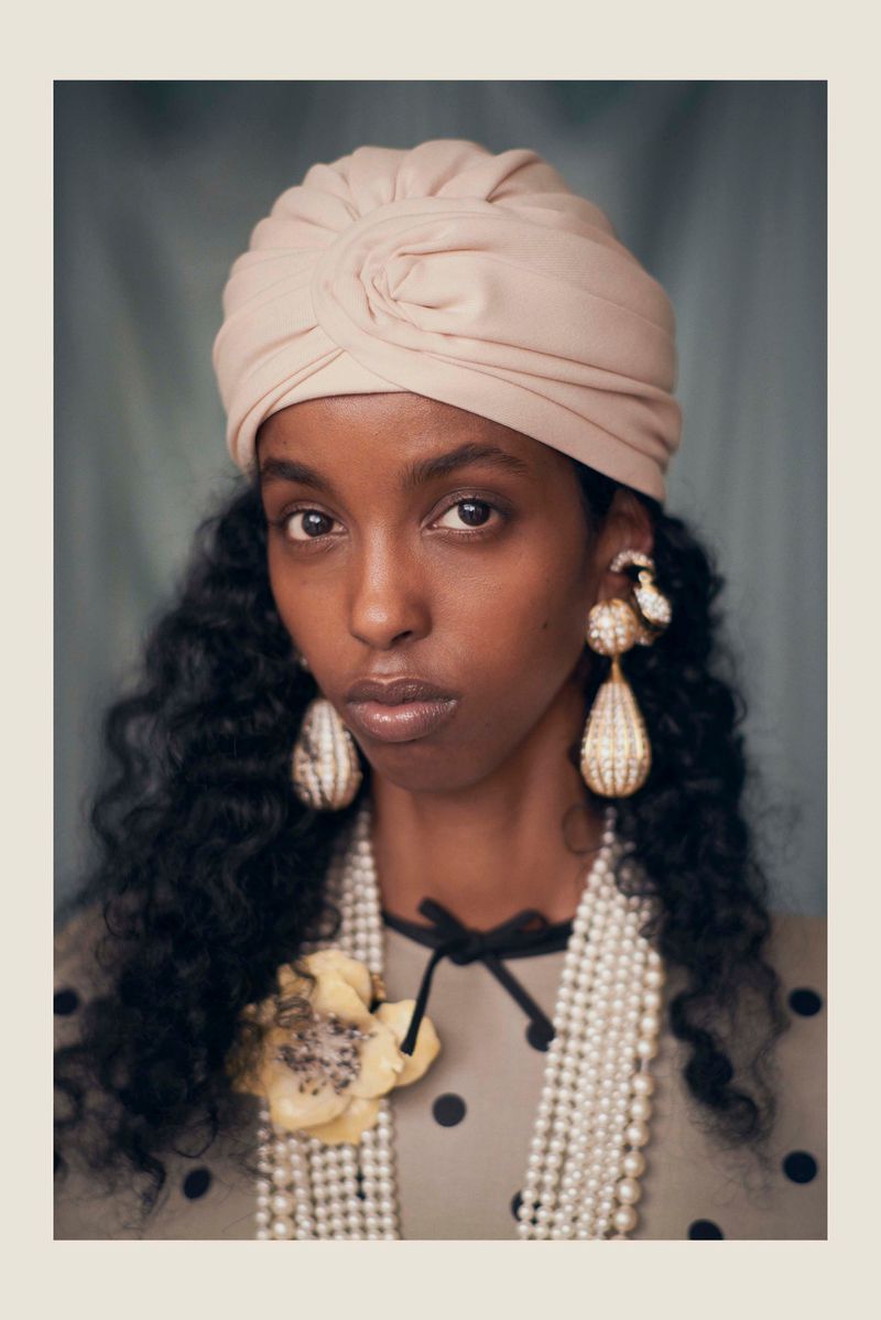 A dark-skinned woman with curly hair wears a beige turban, pearl necklaces, and earrings in a portrait with a muted-green backdrop.