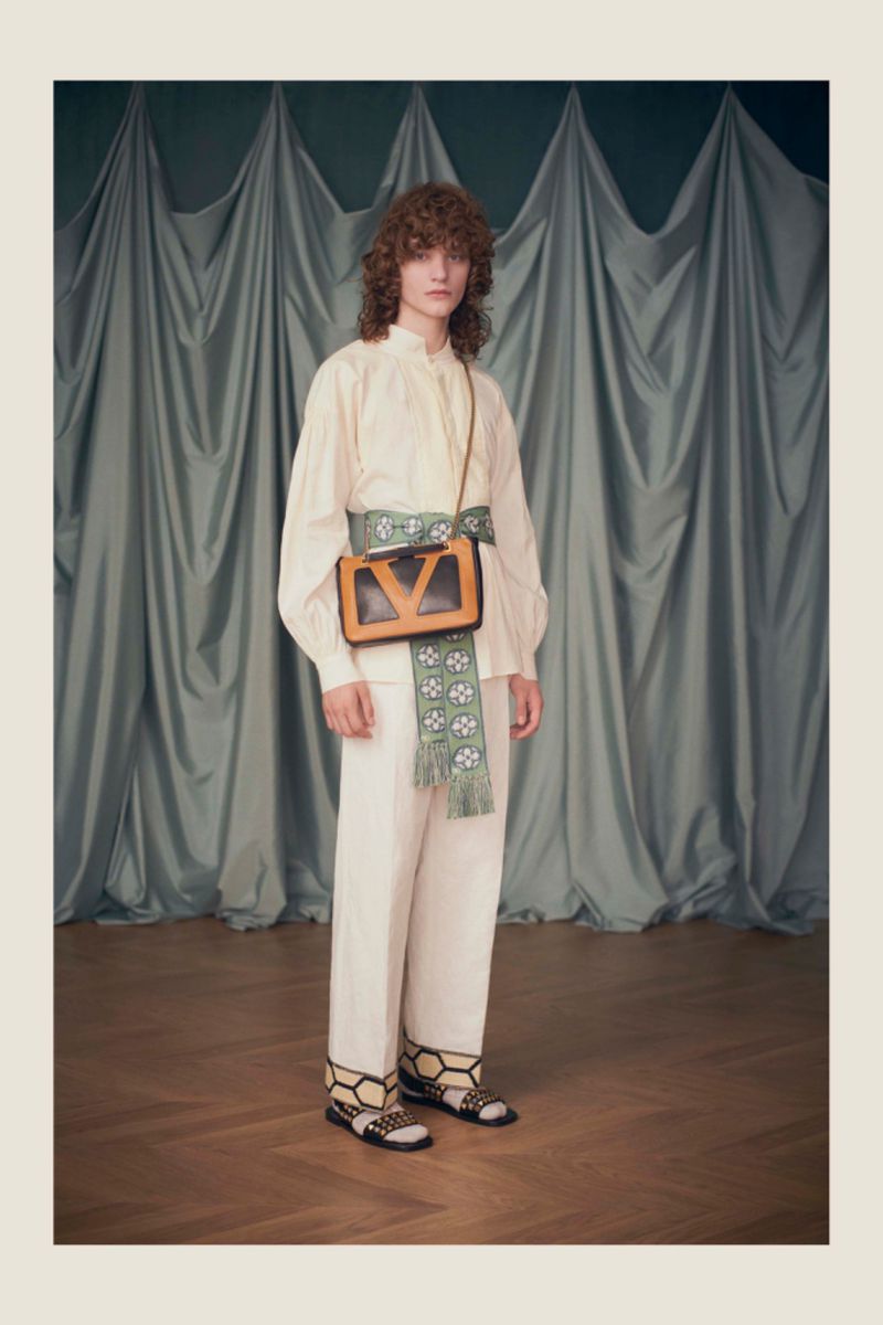 Full shot of a slender adult fashion model wearing an off-white blouse and pants, bag and sandals in a photo studio.