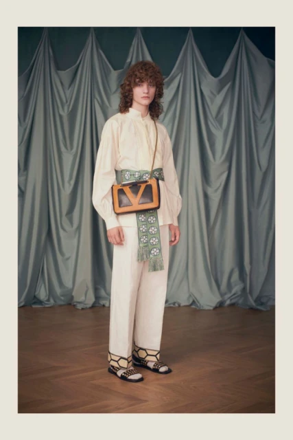 Full shot of a slender adult fashion model wearing an off-white blouse and pants, bag and sandals in a photo studio.
