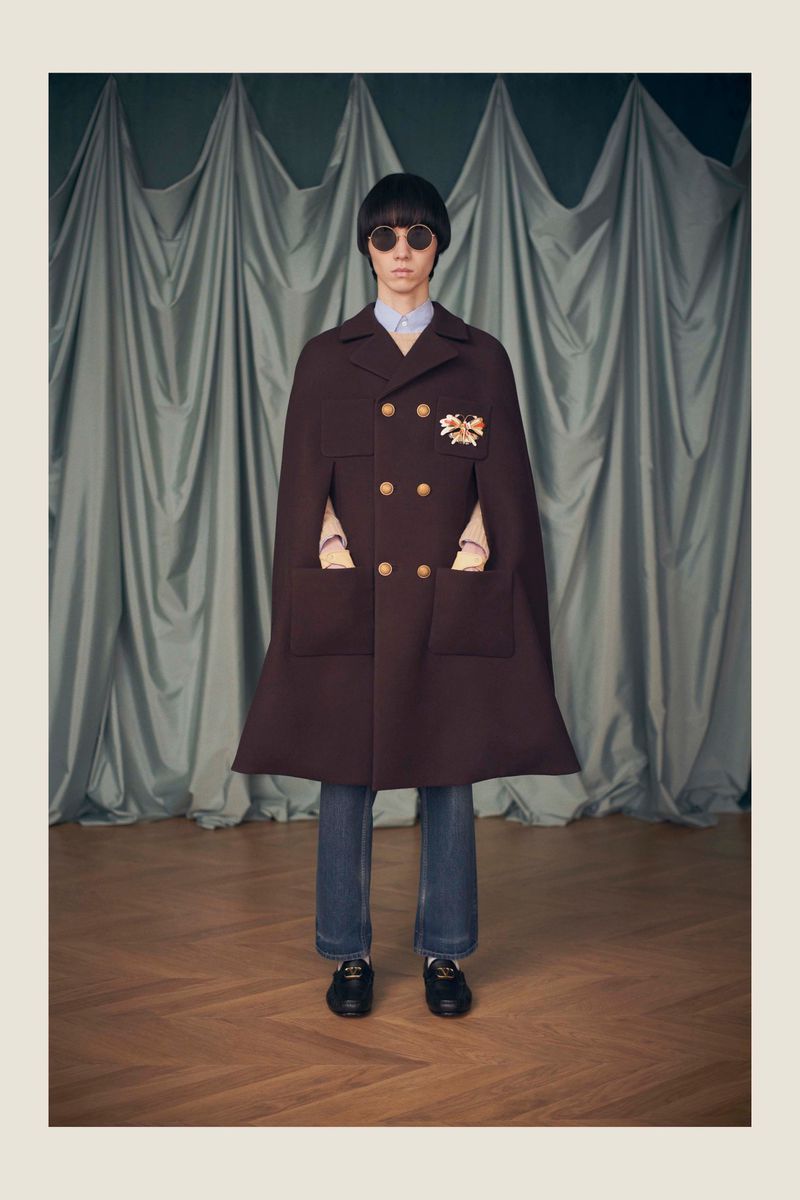 Adult posing in a brown cape-coat with glasses, blue jeans, and loafers, standing in front of a draped grey curtain and wood floor.