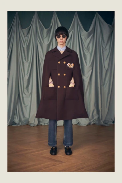 Adult posing in a brown cape-coat with glasses, blue jeans, and loafers, standing in front of a draped grey curtain and wood floor.