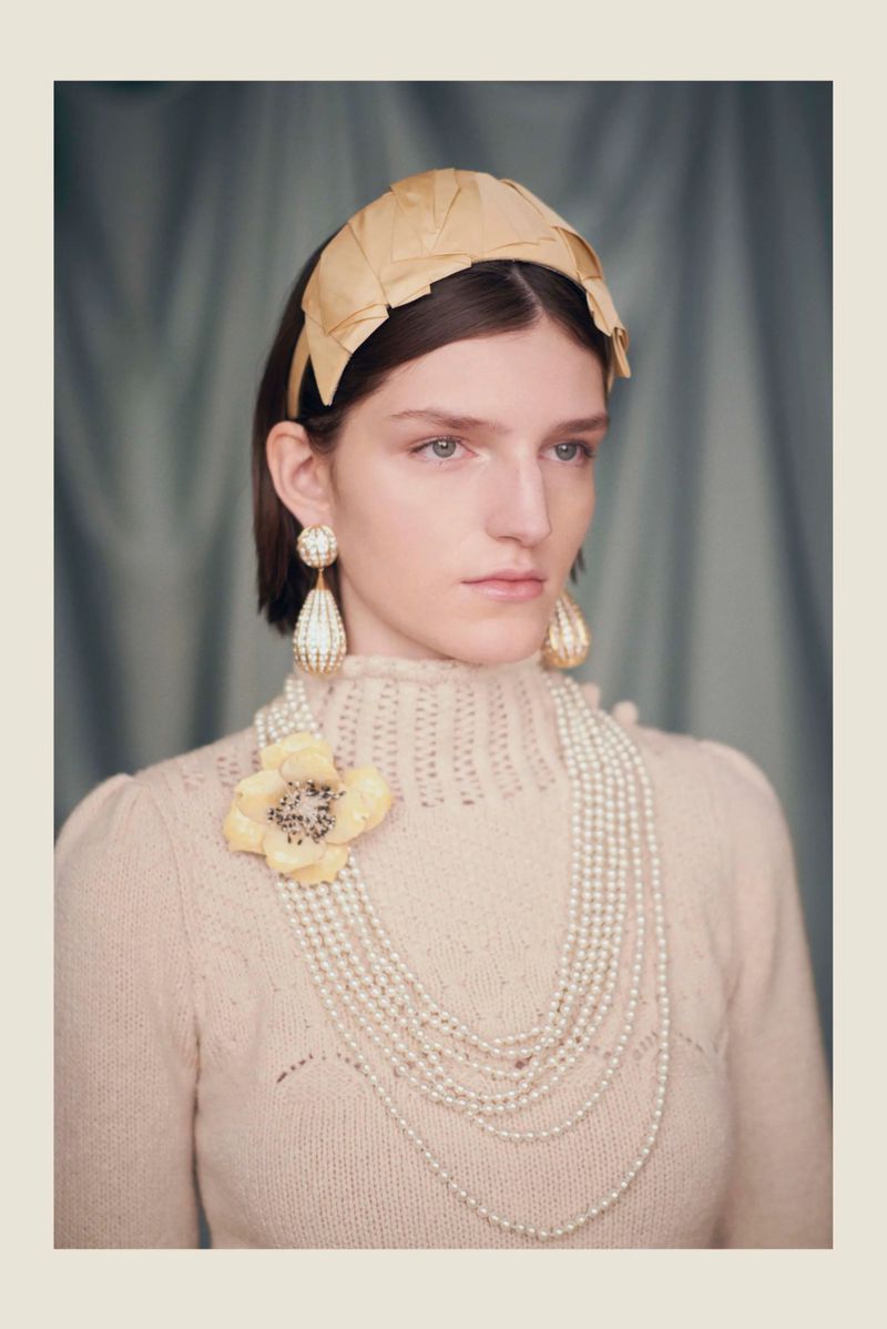 Close-up shot of a woman wearing a cream sweater, pearl jewelry, and a woven headband against a muted blue background.