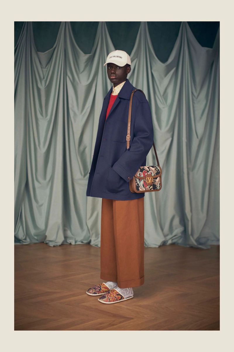 Studio shot of a black male model in a navy jacket, wide-leg trousers, slides, and a crossbody bag against a draped backdrop.