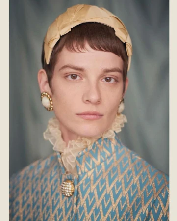 Close-up portrait of a model wearing a yellow pleated headband, ruffled collar, and blue patterned jacket.