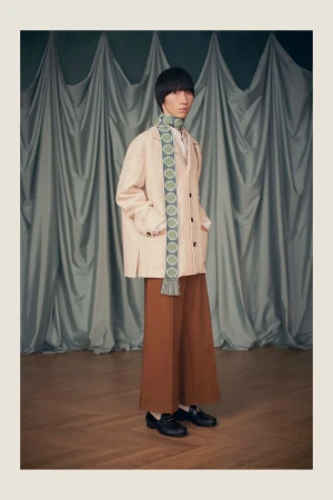 Model wearing a beige tailored jacket, patterned scarf, and wide-leg brown trousers in front of draped curtains on a wood floor.