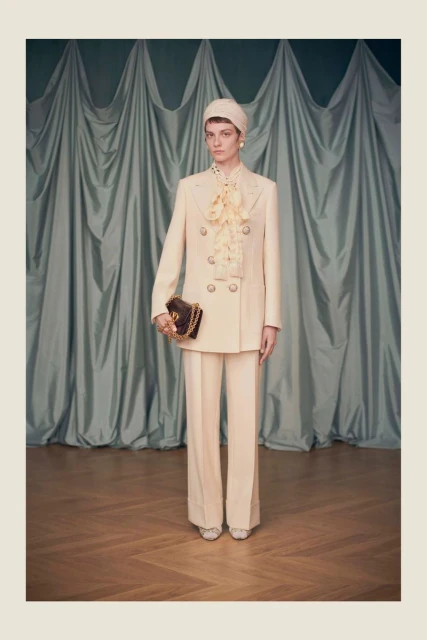 Fashion model wears a cream pantsuit with a ribboned blouse, hat, and purse, posing in front of a pale teal curtain backdrop.