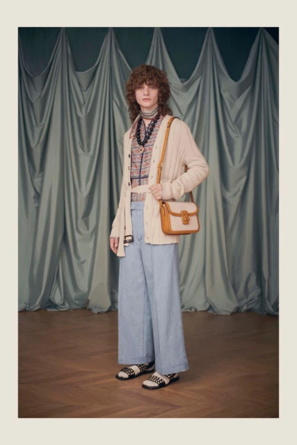 A slender person with shoulder-length curly hair wears an open tan cardigan, patterned top, light blue wide leg jeans and sandals.