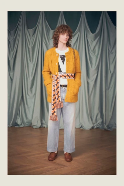 Full-length shot of a person with curly hair in a mustard cardigan, white tee, and light wash jeans, styled with a patterned belt.