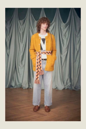 Full-length shot of a person with curly hair in a mustard cardigan, white tee, and light wash jeans, styled with a patterned belt.