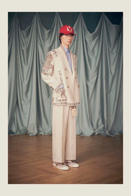 Male model poses in a white double-breasted suit and red cap against a draped blue curtain backdrop, showcasing high fashion.