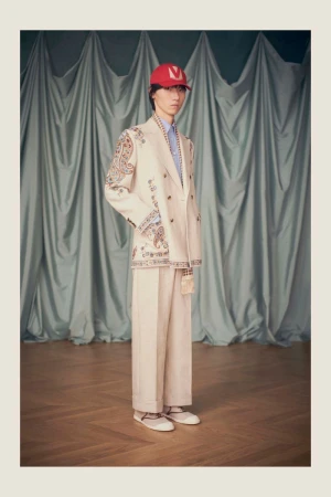 Male model poses in a white double-breasted suit and red cap against a draped blue curtain backdrop, showcasing high fashion.