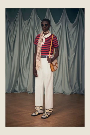 An adult model poses indoors in a red and navy striped shirt, white slacks, round sunglasses, and sandals, with a background of light-green fabric.