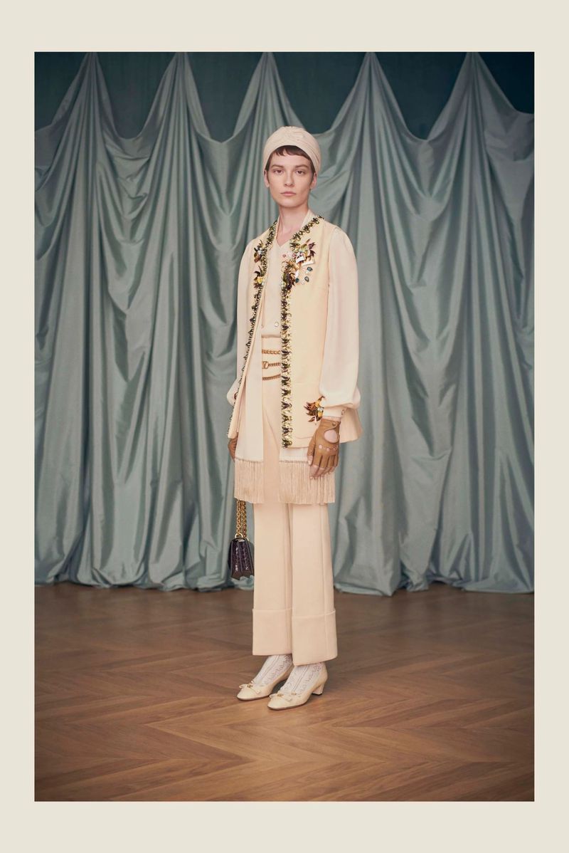 A woman in a cream-colored outfit stands in front of a curtain backdrop with floral embroidery details on a vest and purse.