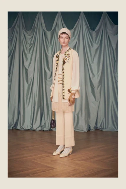 A woman in a cream-colored outfit stands in front of a curtain backdrop with floral embroidery details on a vest and purse.