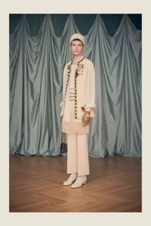 A woman in a cream-colored outfit stands in front of a curtain backdrop with floral embroidery details on a vest and purse.