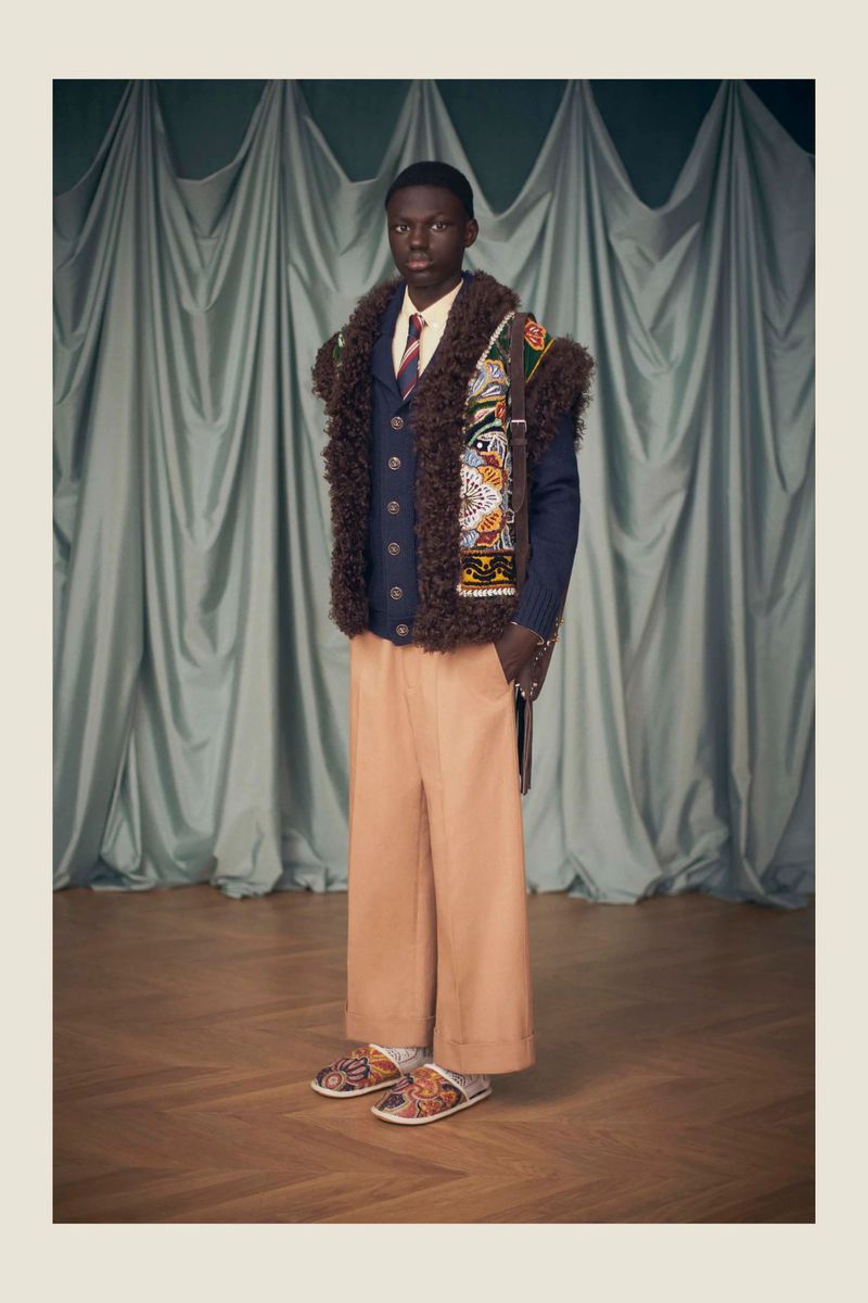 Studio shot of a Black male model in Gucci fashion, wearing a knit cardigan with fur trim, beige pants, and patterned slide sandals.