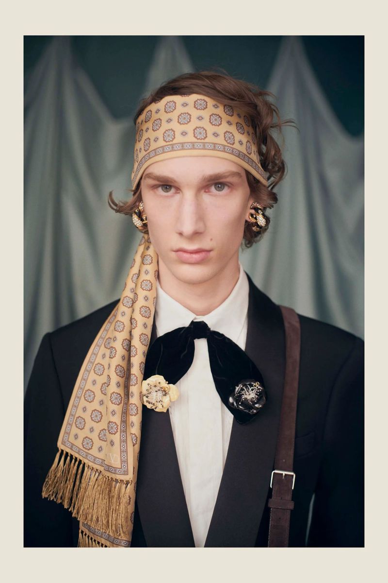 Elegant studio portrait of a fair-skinned adult male wearing patterned headscarf, black suit, and decorative velvet bow tie against a teal backdrop.