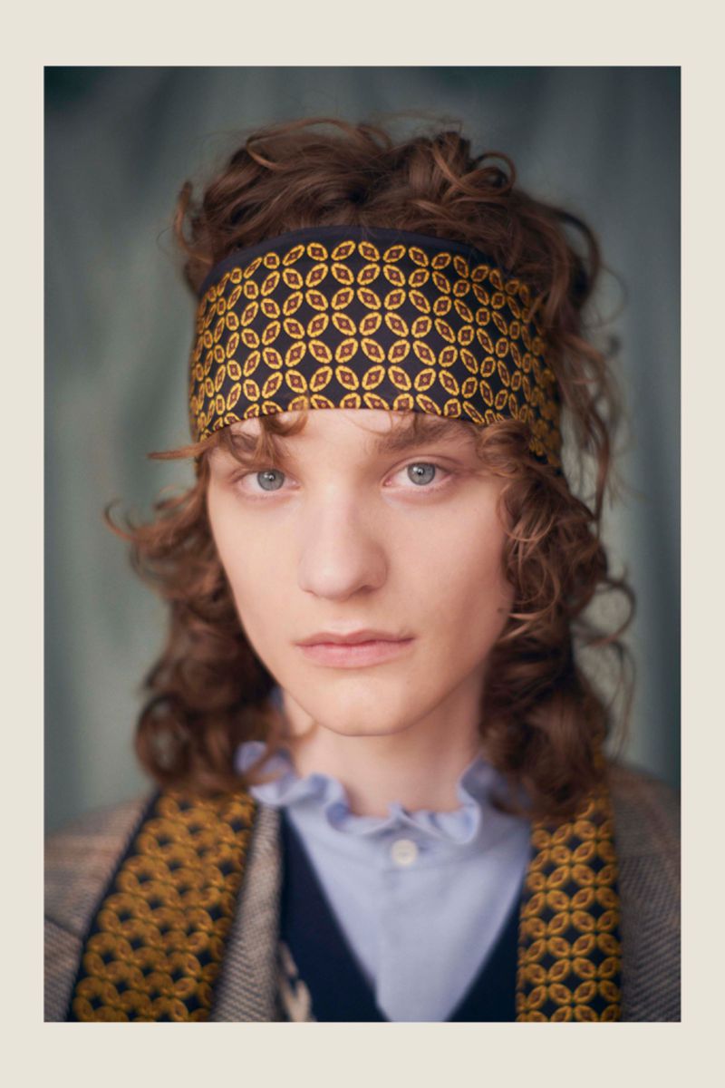 Close-up portrait of a young adult with light skin, curly brown hair, and blue eyes, wearing a patterned headband and a layered outfit.