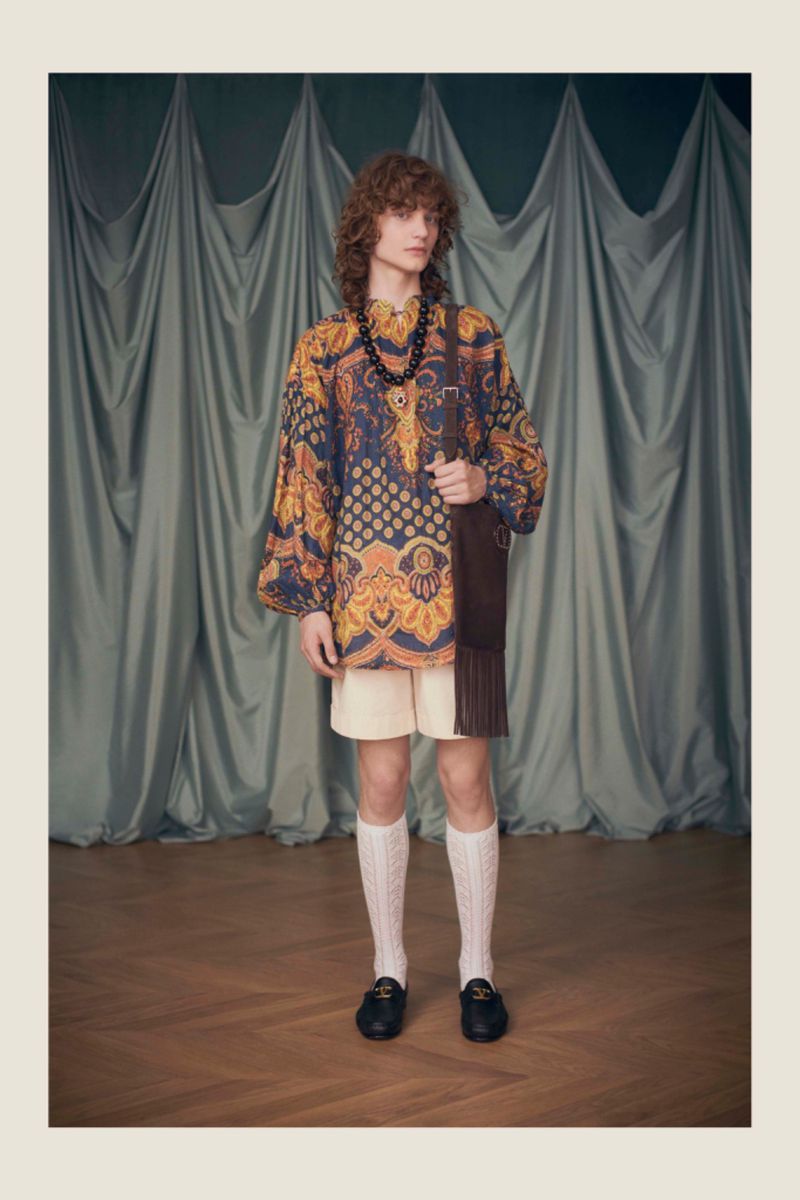 A male model stands in a studio wearing an ornate patterned tunic, shorts, cable knit socks, loafers, and a brown fringed bag.