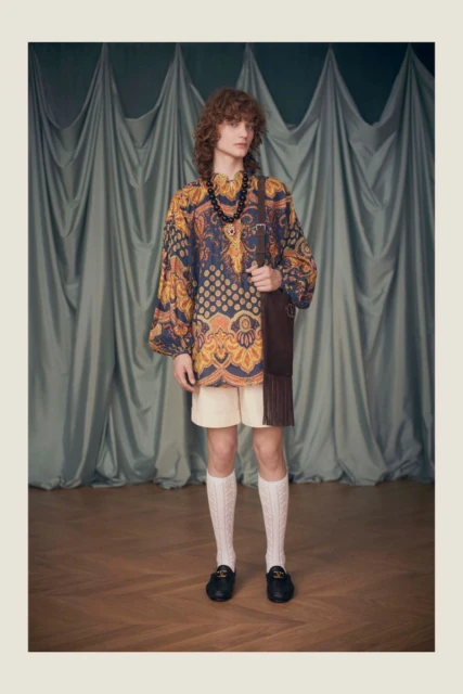 A male model stands in a studio wearing an ornate patterned tunic, shorts, cable knit socks, loafers, and a brown fringed bag.
