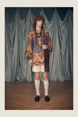 A male model stands in a studio wearing an ornate patterned tunic, shorts, cable knit socks, loafers, and a brown fringed bag.