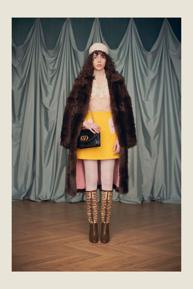 Model wearing a brown fur coat, yellow mini skirt, and knit beanie holding a black shoulder bag in front of draped curtains.
