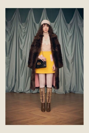Model wearing a brown fur coat, yellow mini skirt, and knit beanie holding a black shoulder bag in front of draped curtains.