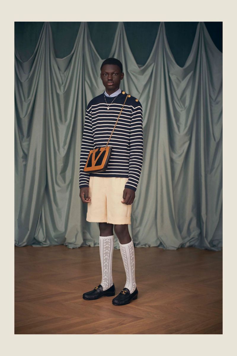 A stylish, full-length studio shot of an African-American man in a striped sweater, shorts, patterned socks, and loafers with a shoulder bag.