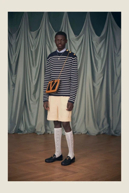 A stylish, full-length studio shot of an African-American man in a striped sweater, shorts, patterned socks, and loafers with a shoulder bag.