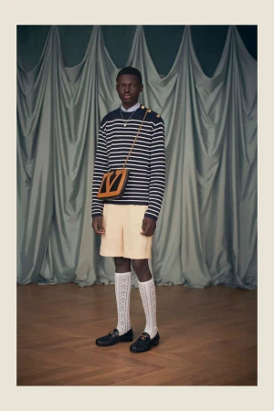 A stylish, full-length studio shot of an African-American man in a striped sweater, shorts, patterned socks, and loafers with a shoulder bag.