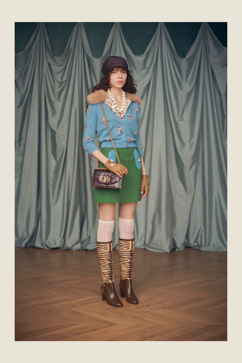 Model posing in a blue cardigan with fur collar, green mini skirt, and patterned tall boots against draped fabric.