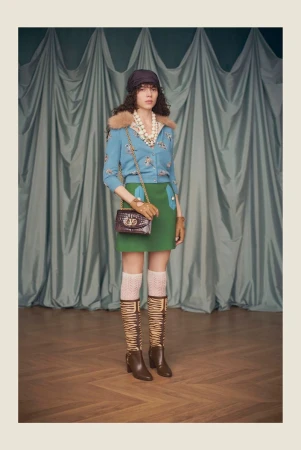 Model posing in a blue cardigan with fur collar, green mini skirt, and patterned tall boots against draped fabric.