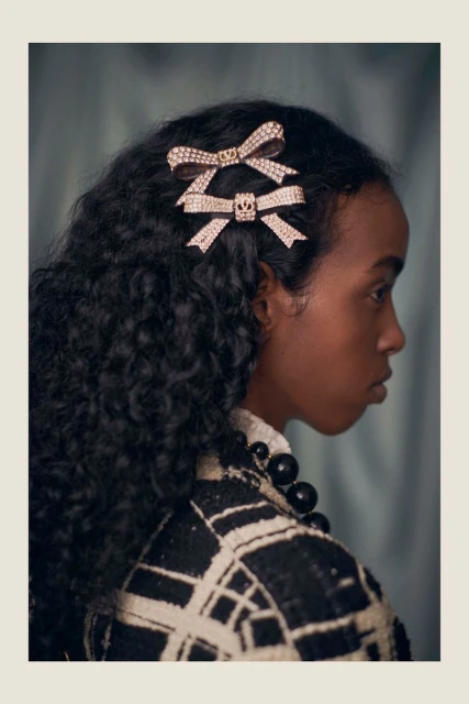 Close-up of a woman with dark curly hair, wearing bejeweled hair clips, a patterned coat, and a large beaded necklace against a muted backdrop.
